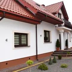 Biały dom z brązowymi rynnami i dachówką. Przed domem ozdobne krzewy i ścieżka z cokolików.