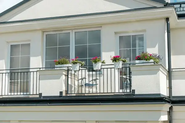 Elewacja domu z balkonem ozdobionym białymi donicami z kwitnącymi kwiatami i czarną, metalową balustradą.